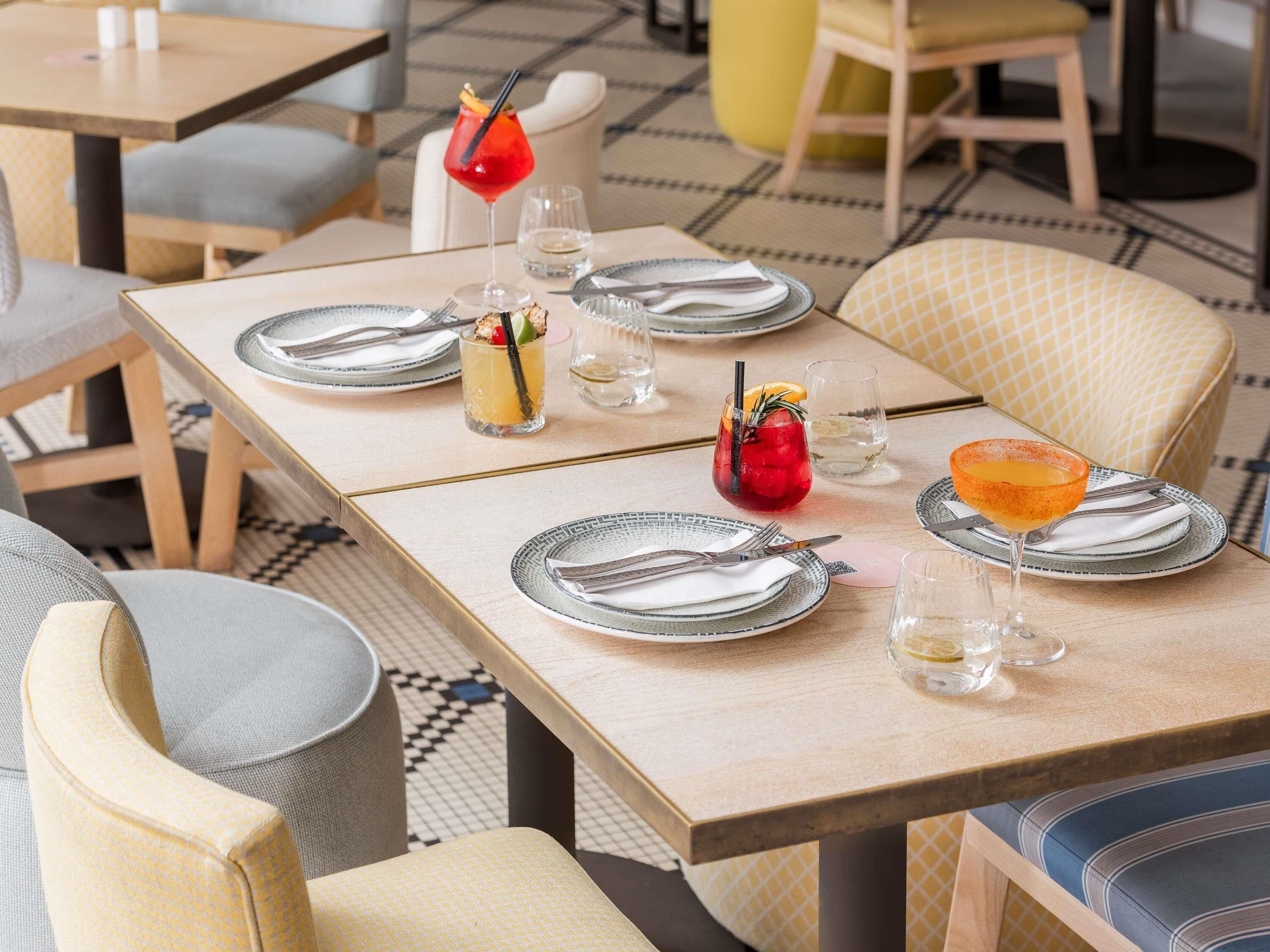 Una mesa de comedor dispuesta con platos, cubiertos y vasos, con un suelo a cuadros y sillas al fondo.