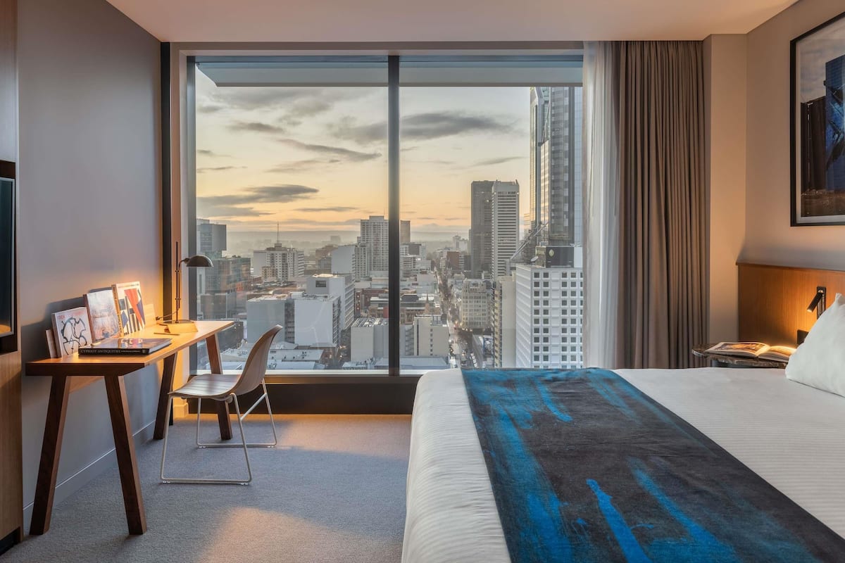 Habitación de hotel con un ventanal grande, un escritorio con silla, una cama y una mesita de noche.