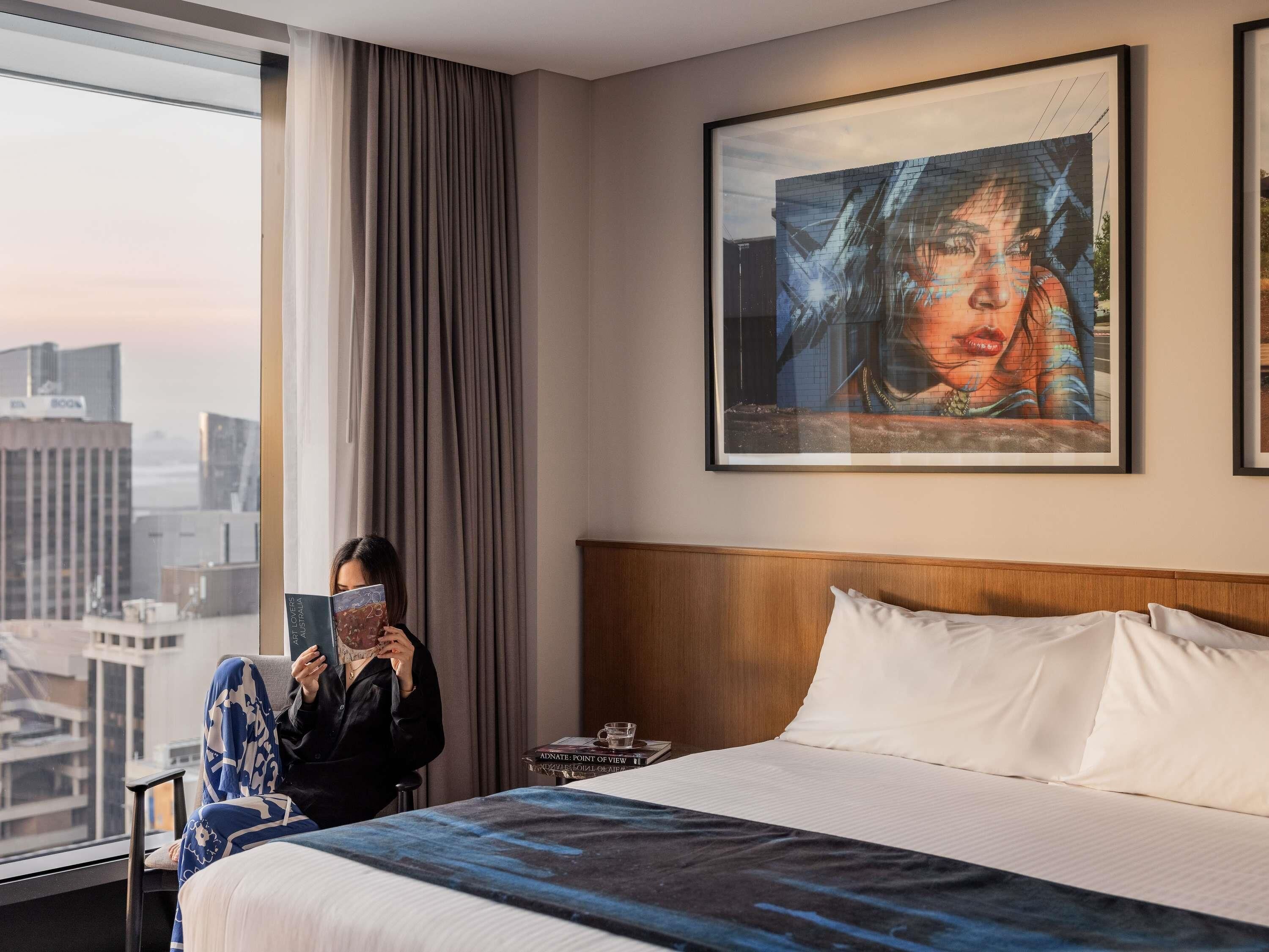 A hotel room with a large bed, a window with curtains, a framed artwork on the wall, and a city view.