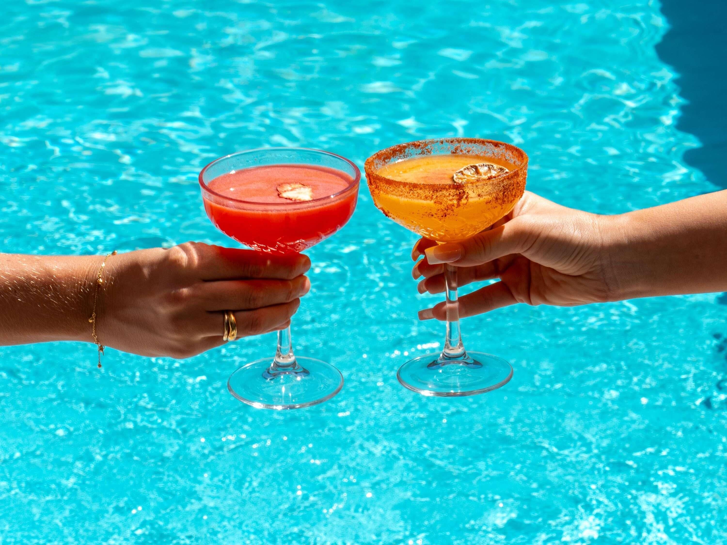 Dos manos sosteniendo copas de cóctel sobre una piscina.