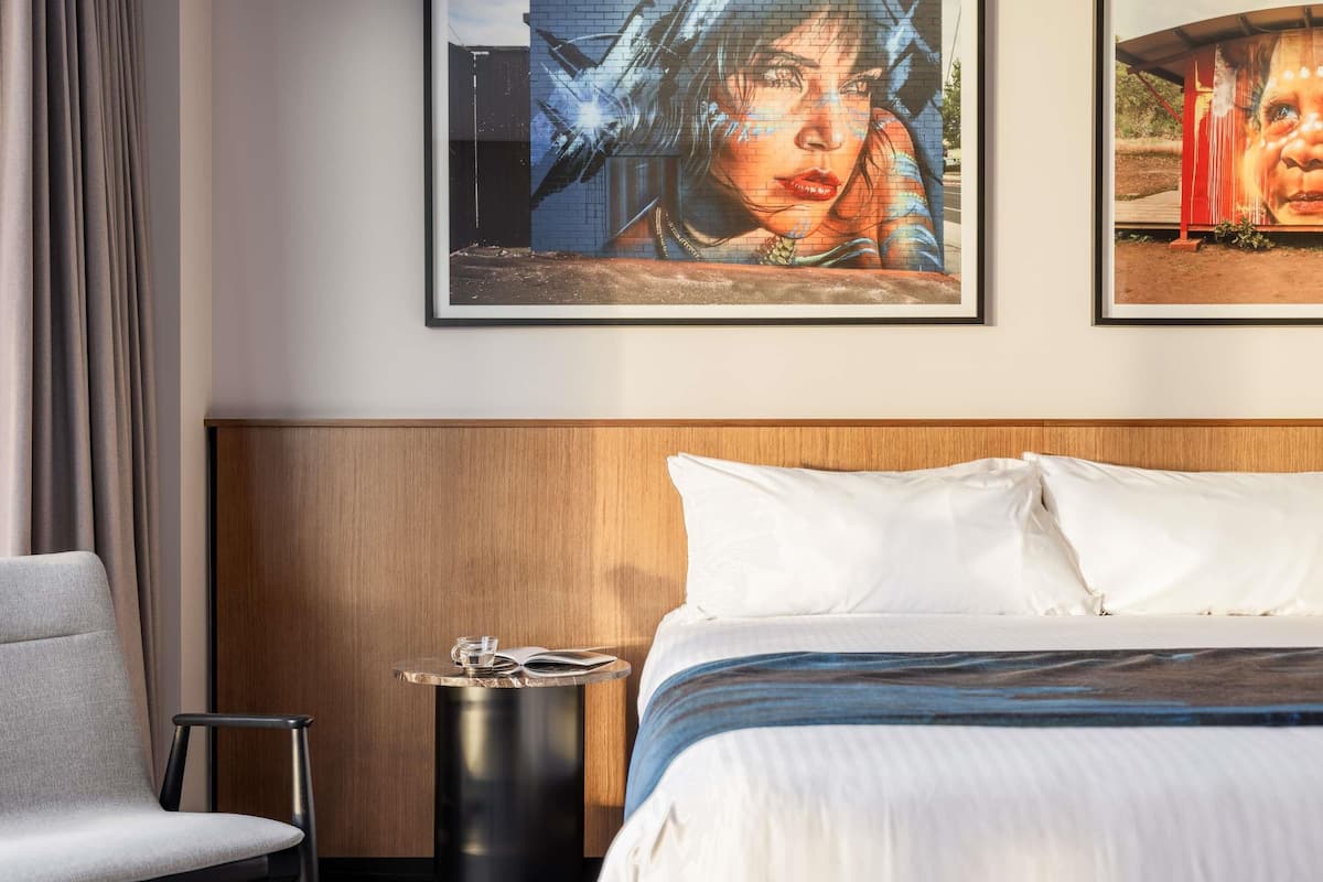 Habitación de hotel con cama, una silla, una mesita y dos cuadros en la pared.