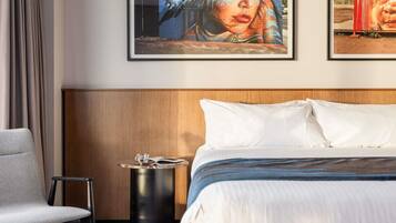 A hotel room with a bed, a chair, a small table, and two framed artworks on the wall.