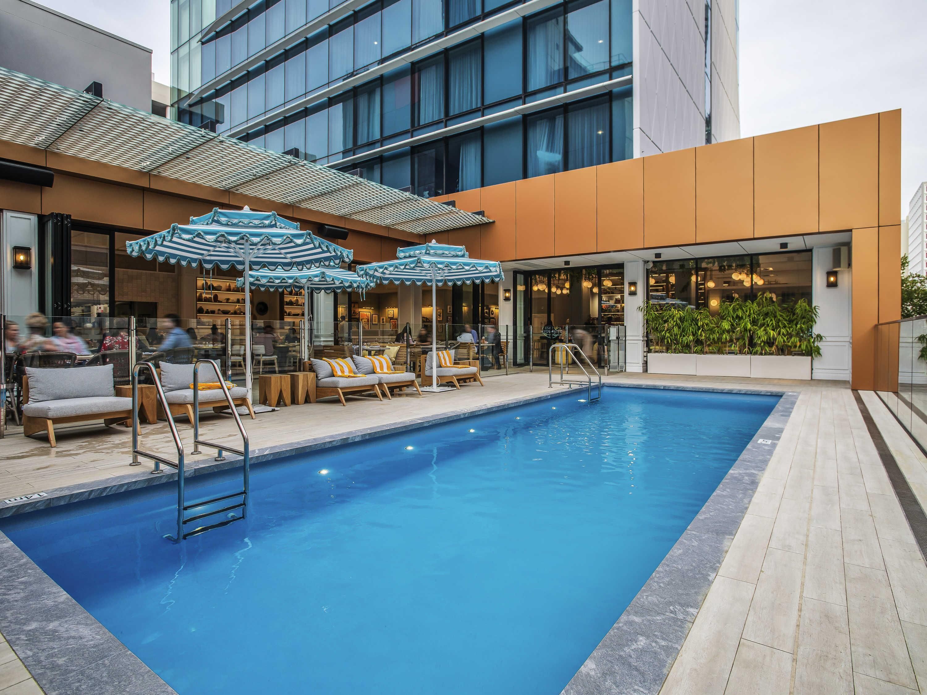 A swimming pool with lounge chairs and umbrellas, a restaurant with indoor seating, and a modern building facade.