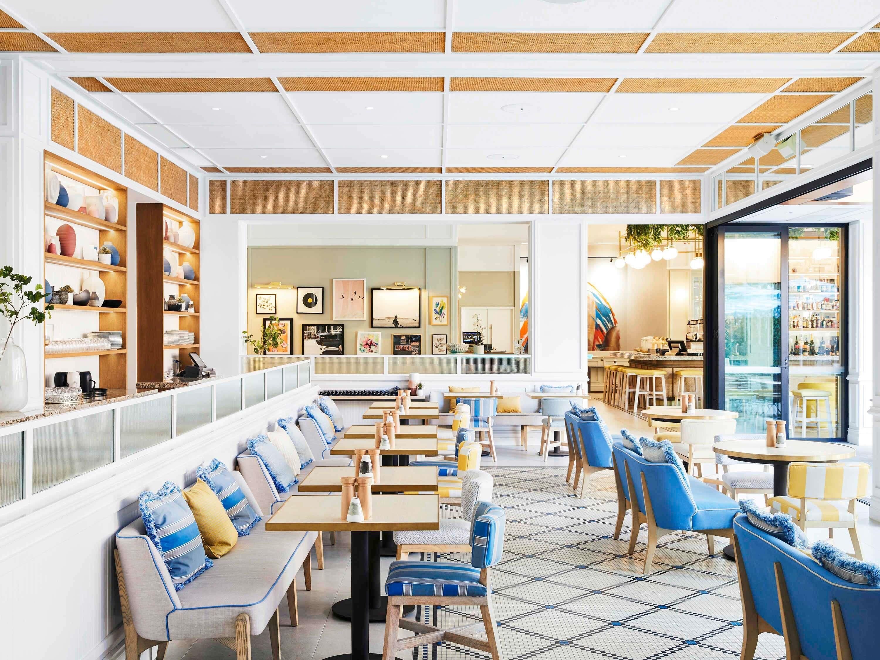 Interior moderno de restaurante con una larga zona de asientos tipo banco, mesas de madera y un área de bar visible a través de un separador de vidrio.