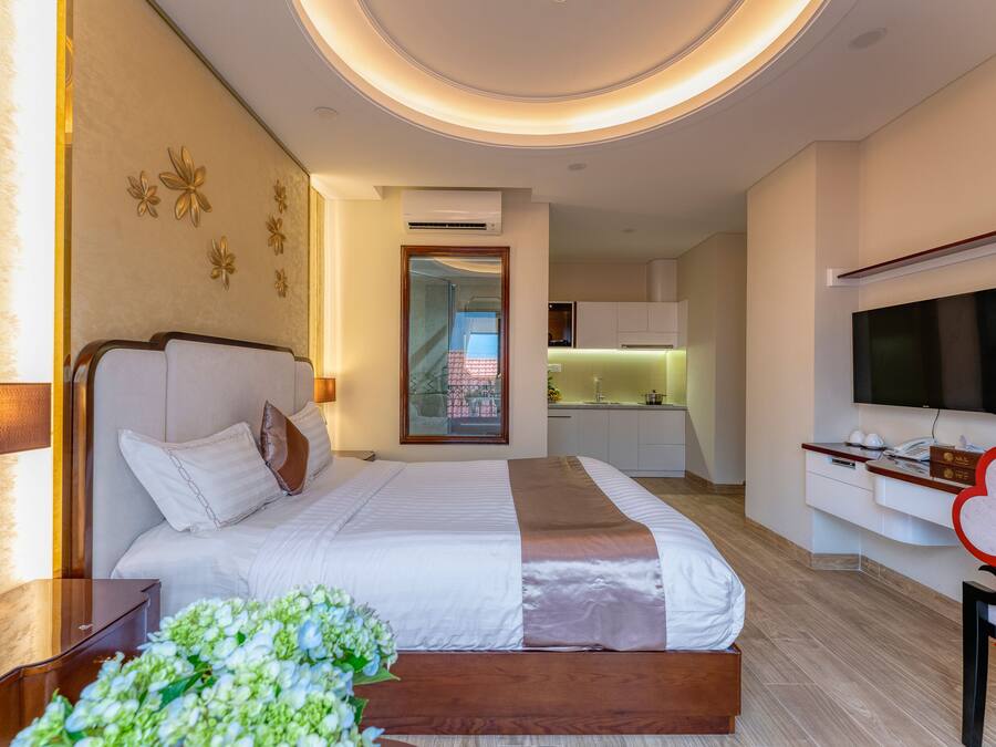 A hotel room with a bed, bedside table, TV, and a view of the kitchen and balcony.