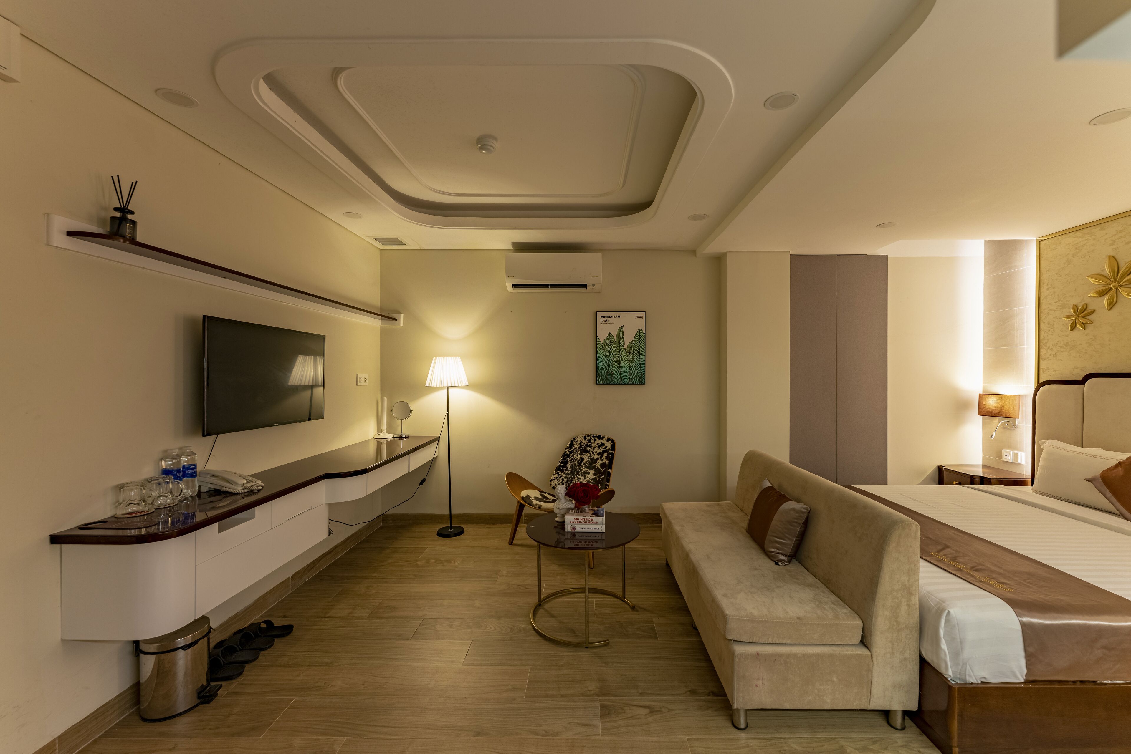 A modern hotel room with a sofa, a bed, a TV, a small table with a lamp, and a trash bin.
