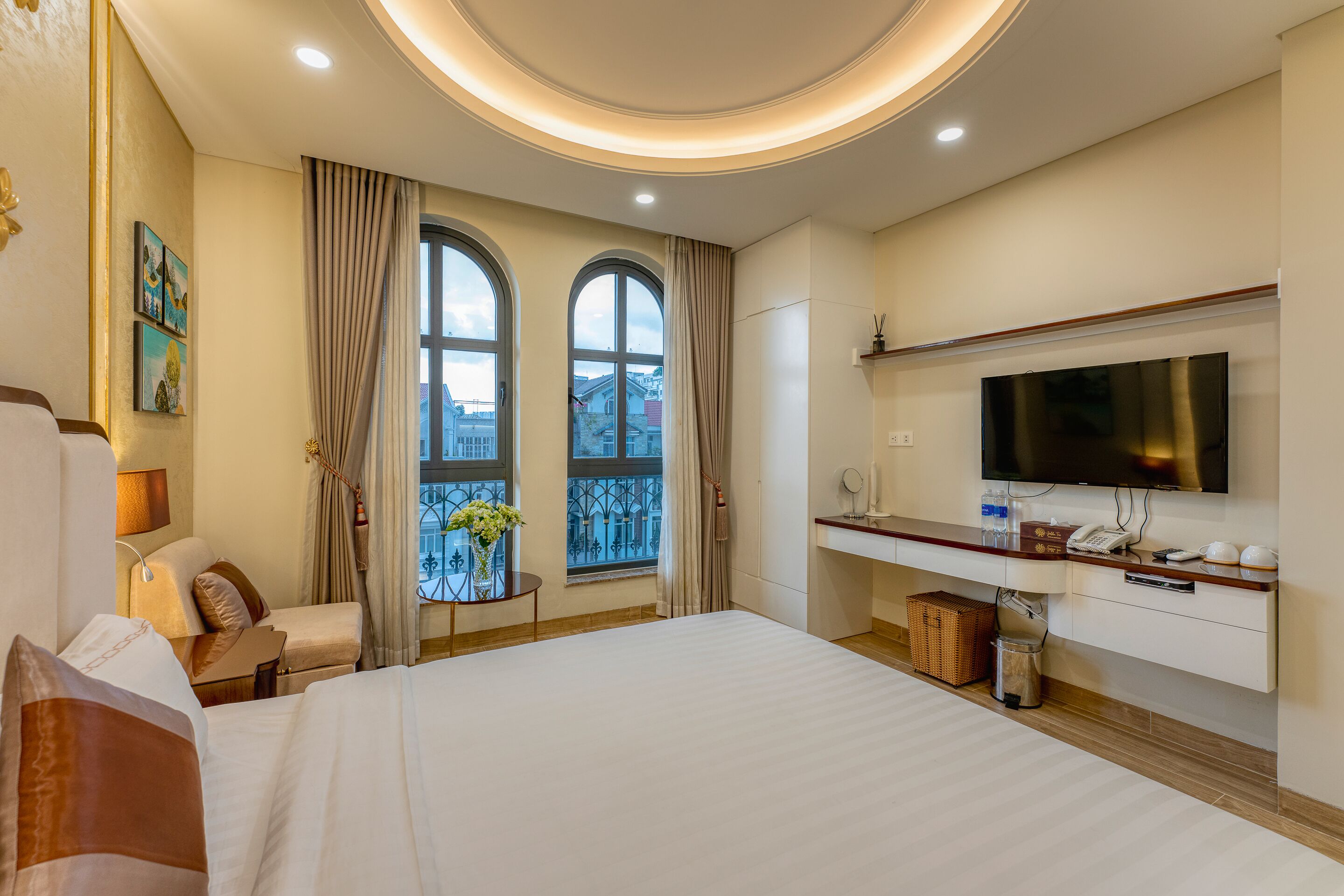 A hotel room with a large bed, a TV, a desk, and a view of the city through two windows.
