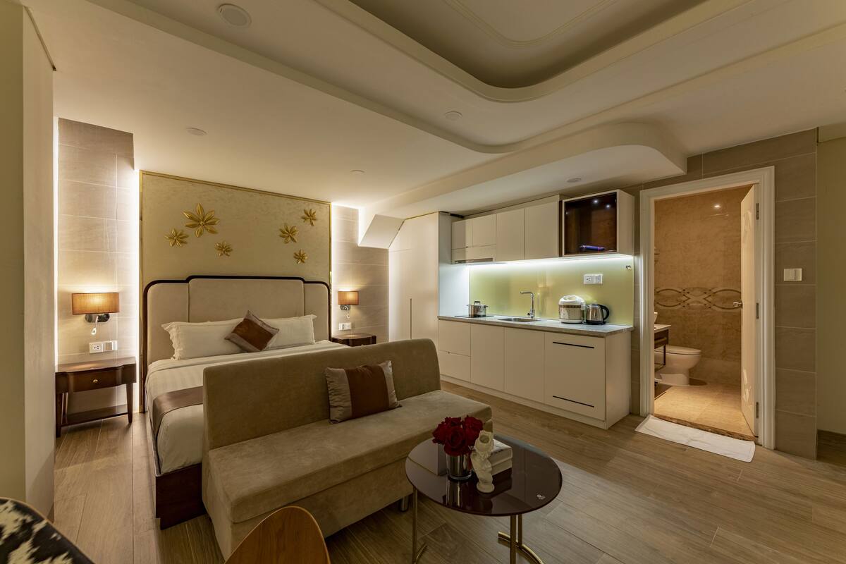 A modern hotel room with a bed, sofa, dining table, and kitchenette.