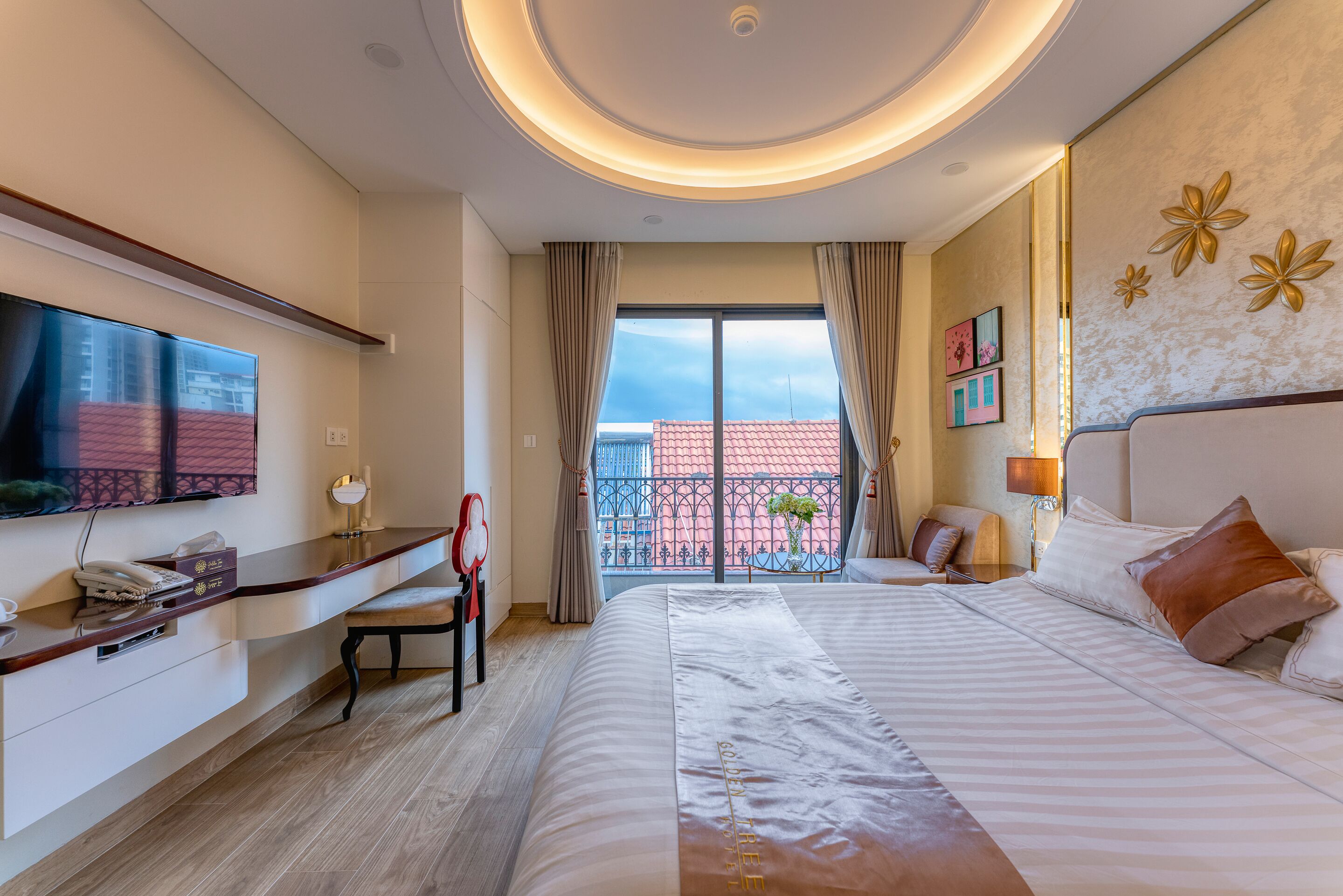 A hotel room with a large bed, a desk with a telephone, a flat-screen TV mounted on the wall, and a balcony with a view of buildings.