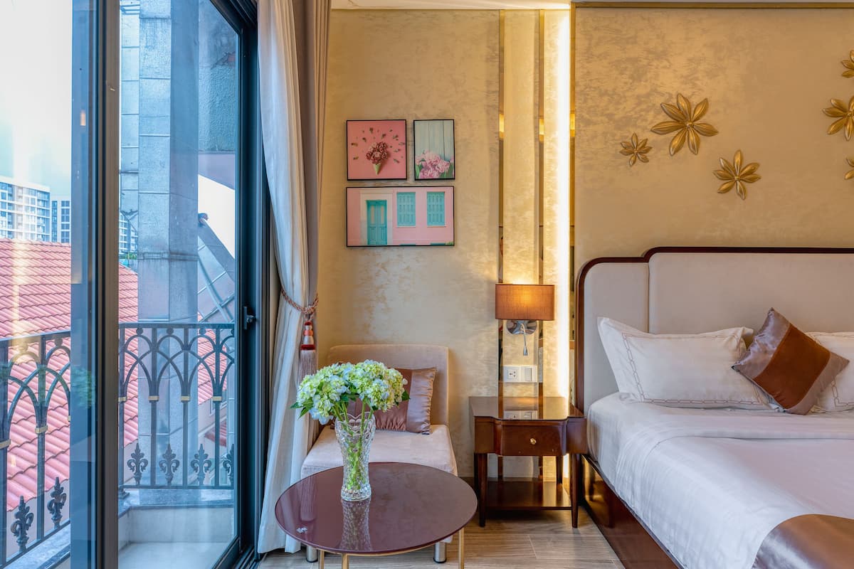 A hotel room with a bed, a bedside table, a lamp, and a vase with flowers. There is a window with a view of buildings outside.
