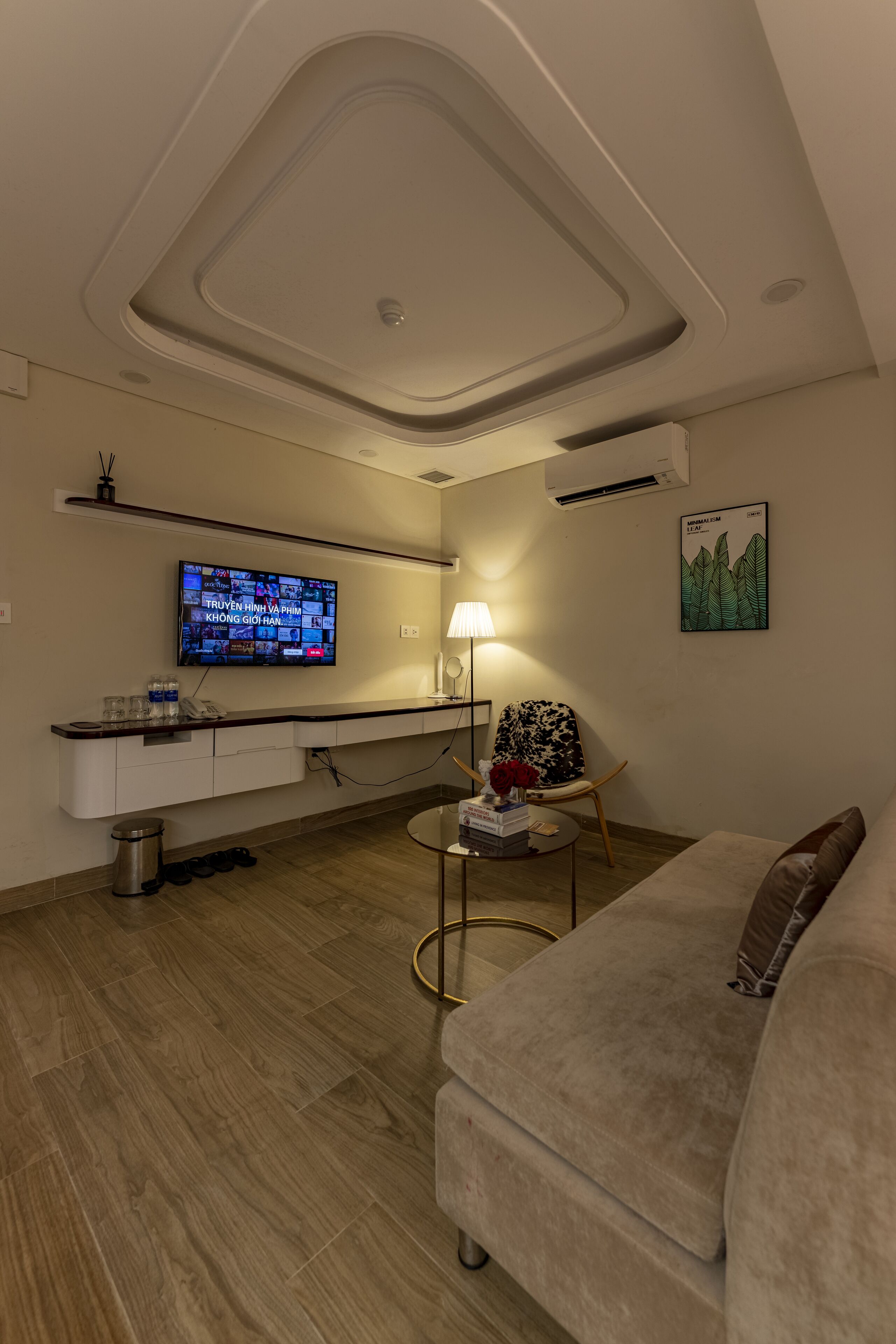 Een moderne woonkamer met een flatscreen televisie aan de muur, een bank, een fauteuil met een gestreept kussen en een klein bijzettafeltje met een lamp en decoratieve objecten.