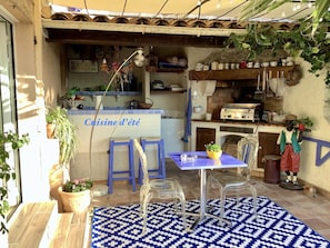 A small outdoor kitchen area with a blue and white patterned rug, two clear plastic chairs, a small table, and various kitchen items on shelves.