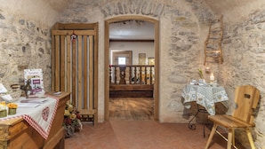 A rustic room with stone walls, a wooden bench, a table with a checkered tablecloth, and a wooden chair. - Horse House (Novella)