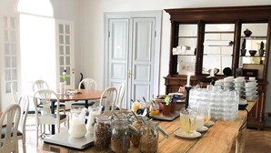 Una zona de menjador amb una taula de fusta llarga, caires blancs i un lampadari. Hi ha pots d'alimentació i una pila de plats sobre la taula.
