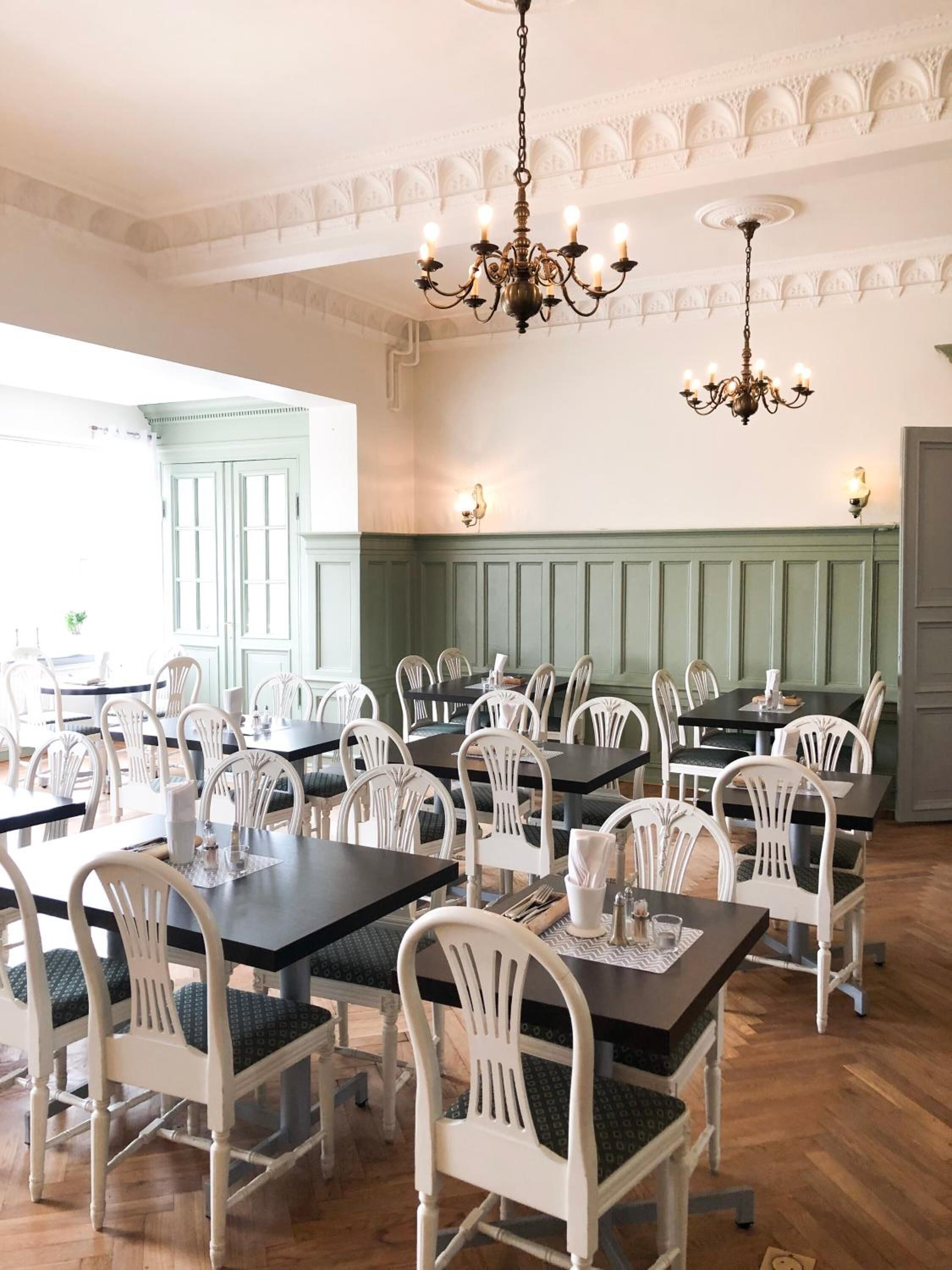 A dining area with white chairs and dark tables, chandeliers, and a green paneled wall.