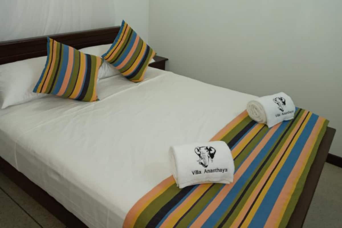 Una cama bien hecha con almohadas y una manta a rayas, una toalla enrollada con un logotipo, y una mesita de noche junto a la cama.
