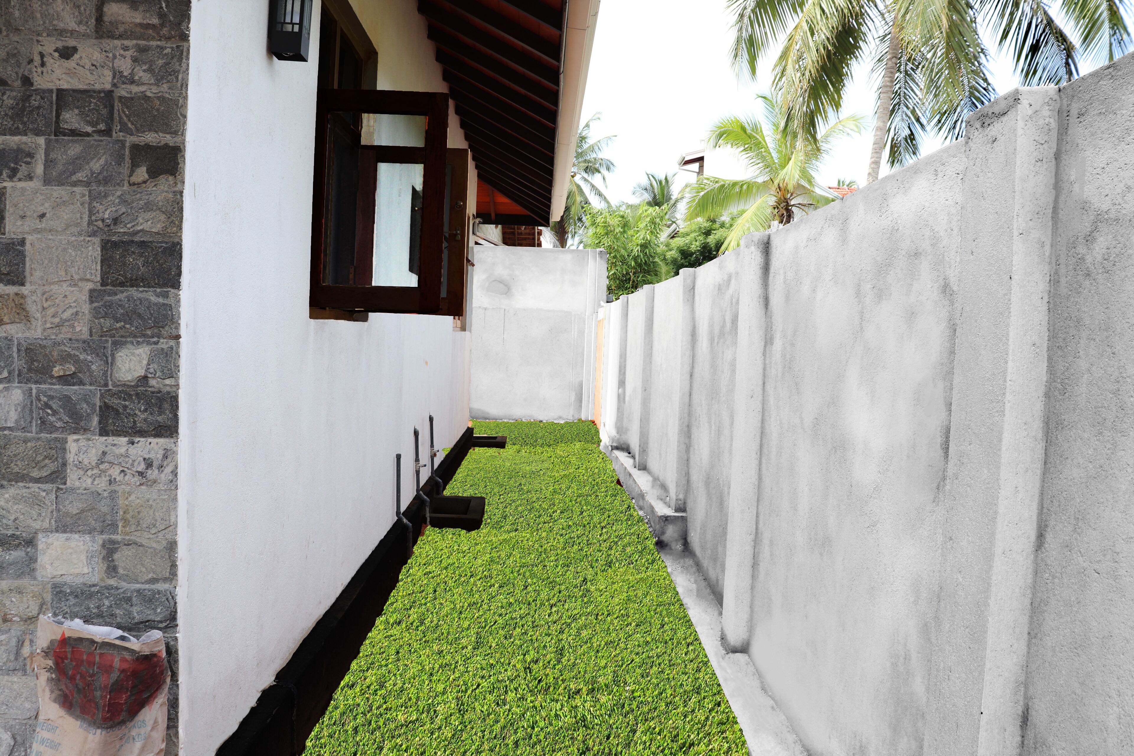 A narrow pathway with artificial green grass, flanked by a stone wall on one side and a concrete wall on the other.