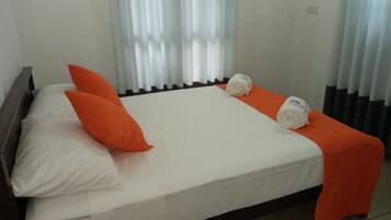 Cama com roupa de cama branca, dois travesseiros laranja e duas toalhas enroladas.