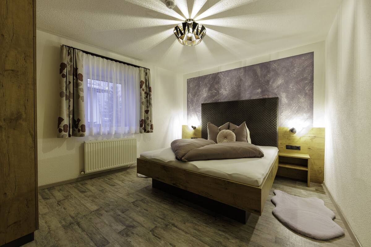 A bedroom with a wooden bed, a radiator, a window with curtains, a wall-mounted light, and a headboard.