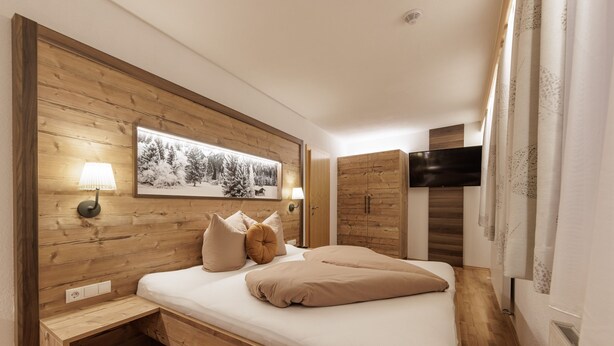 A bedroom with a wooden headboard, a bed with beige bedding, a bedside table, a wall-mounted TV, and a framed picture above the bed. - Apartment Goldeck (Ried im Oberinntal)