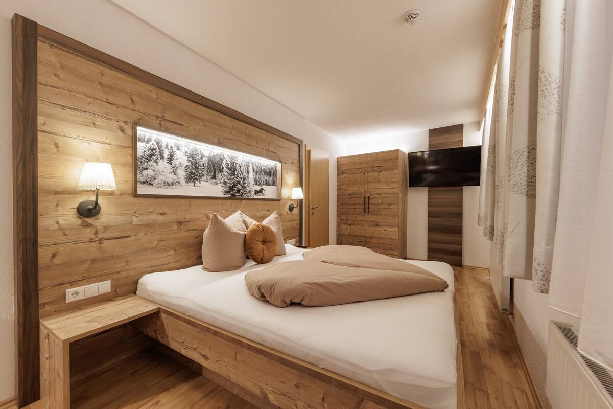 A bedroom with a wooden headboard, a bed with beige bedding, a bedside table, a wall-mounted TV, and a framed picture above the bed.