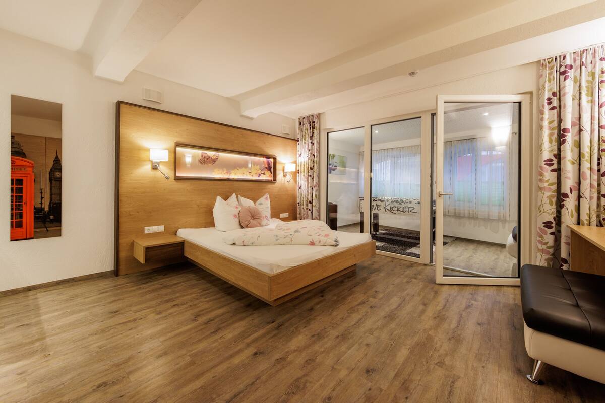 A modern hotel room with a wooden bed, a small bedside table, a wall-mounted light, and a large mirror.