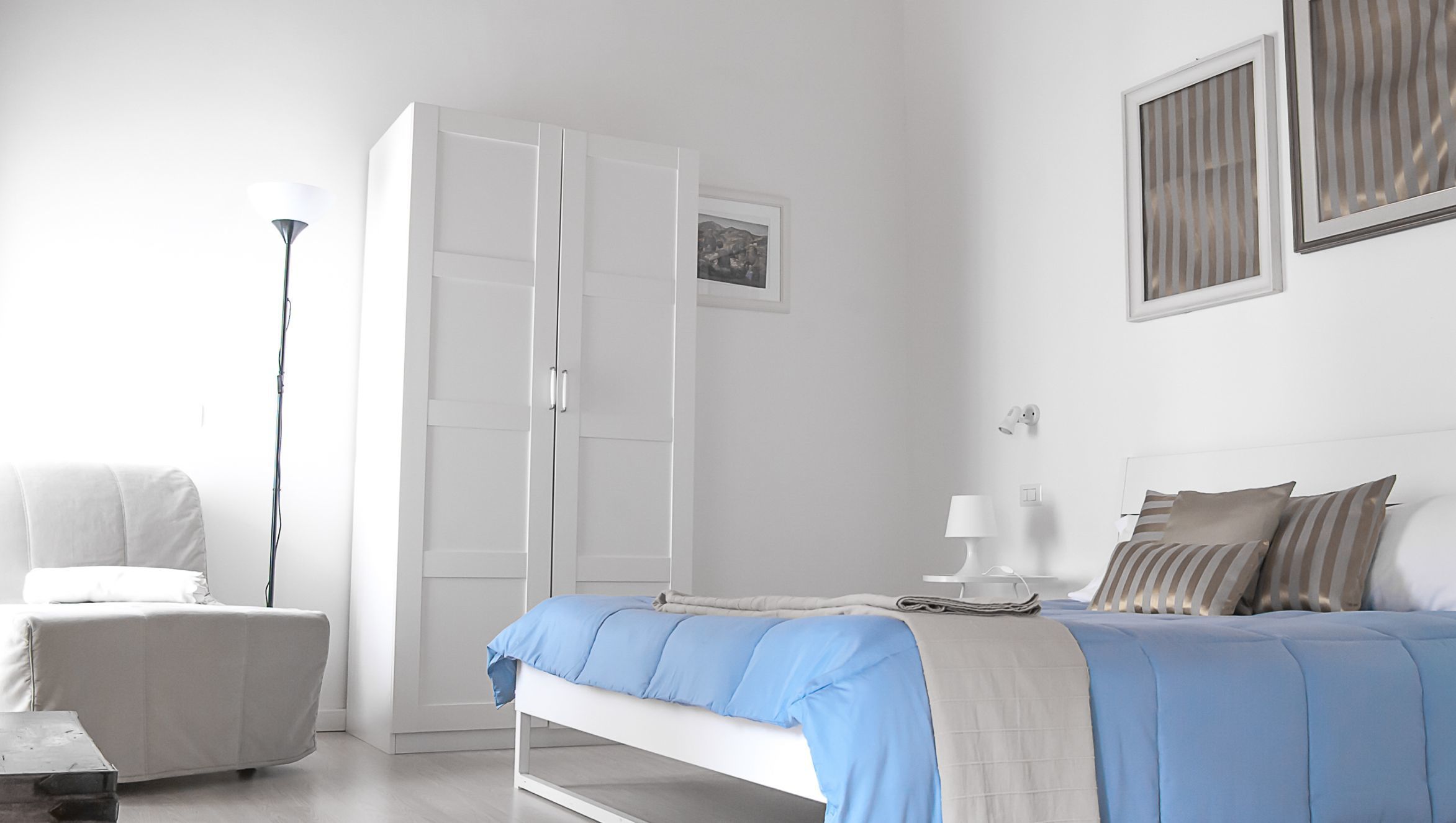 A bedroom with a bed, a chair, a wardrobe, a bedside table with a lamp, and a picture on the wall.