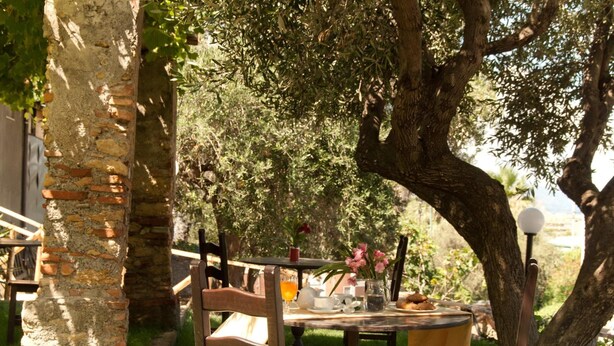 An outdoor dining area with a brick pillar, wooden chairs, and a table set with a pitcher, cups, and a plate of food. - Bookbreakfast (Terme Vigliatore)
