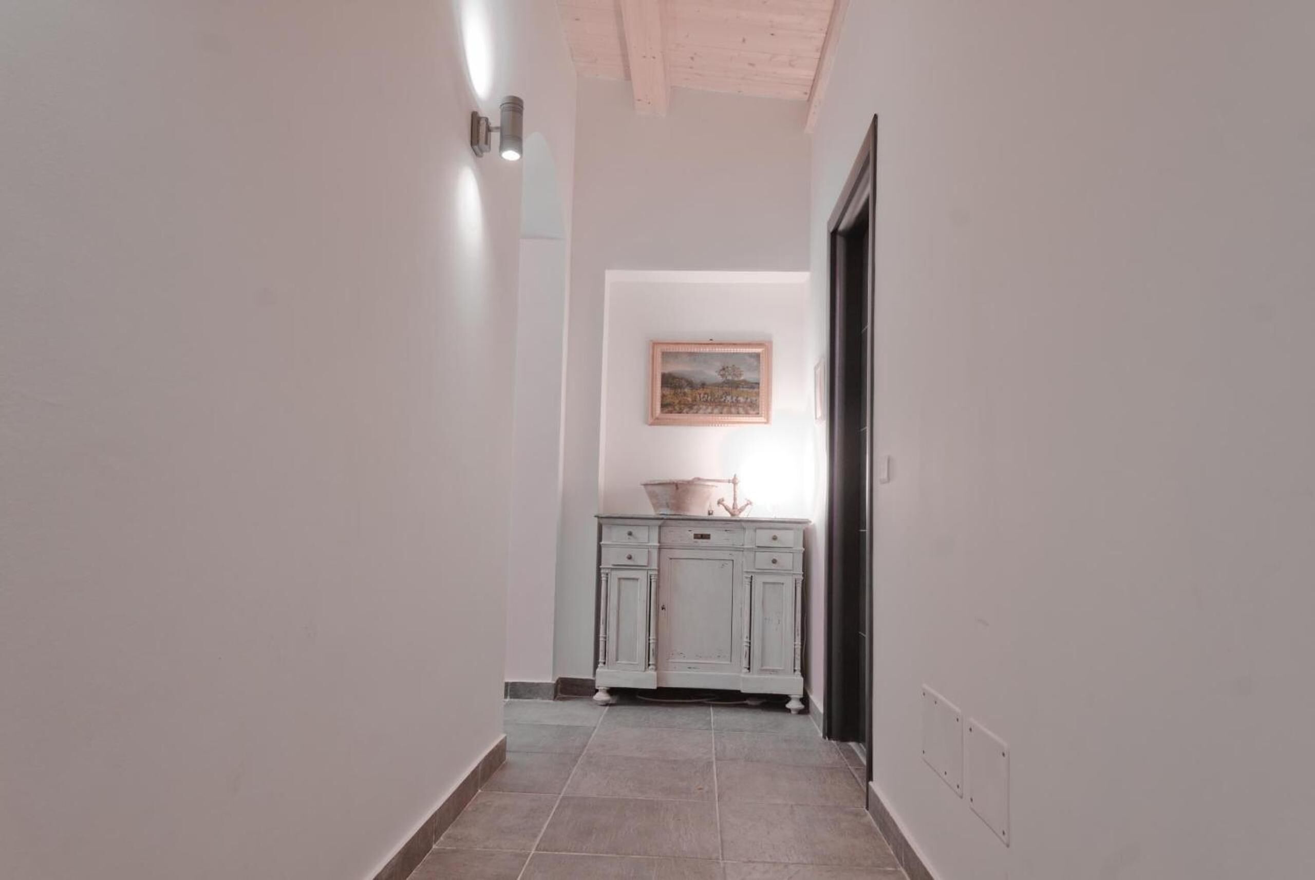 Um corredor estreito com chão de azulejos claros, parede branca com um quadro emoldurado e um armário branco com gavetas.