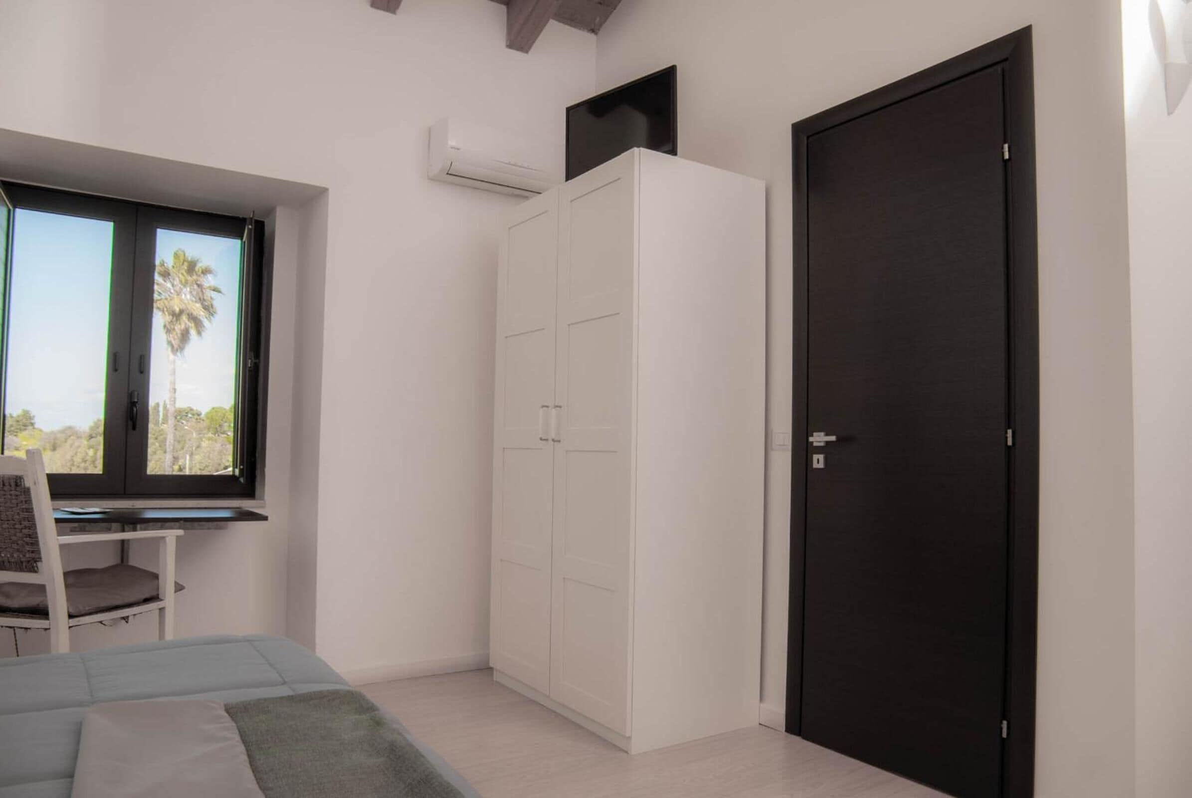 Une chambre à coucher comprenant un lit, un bureau, une armoire et une fenêtre donnant sur des palmiers.