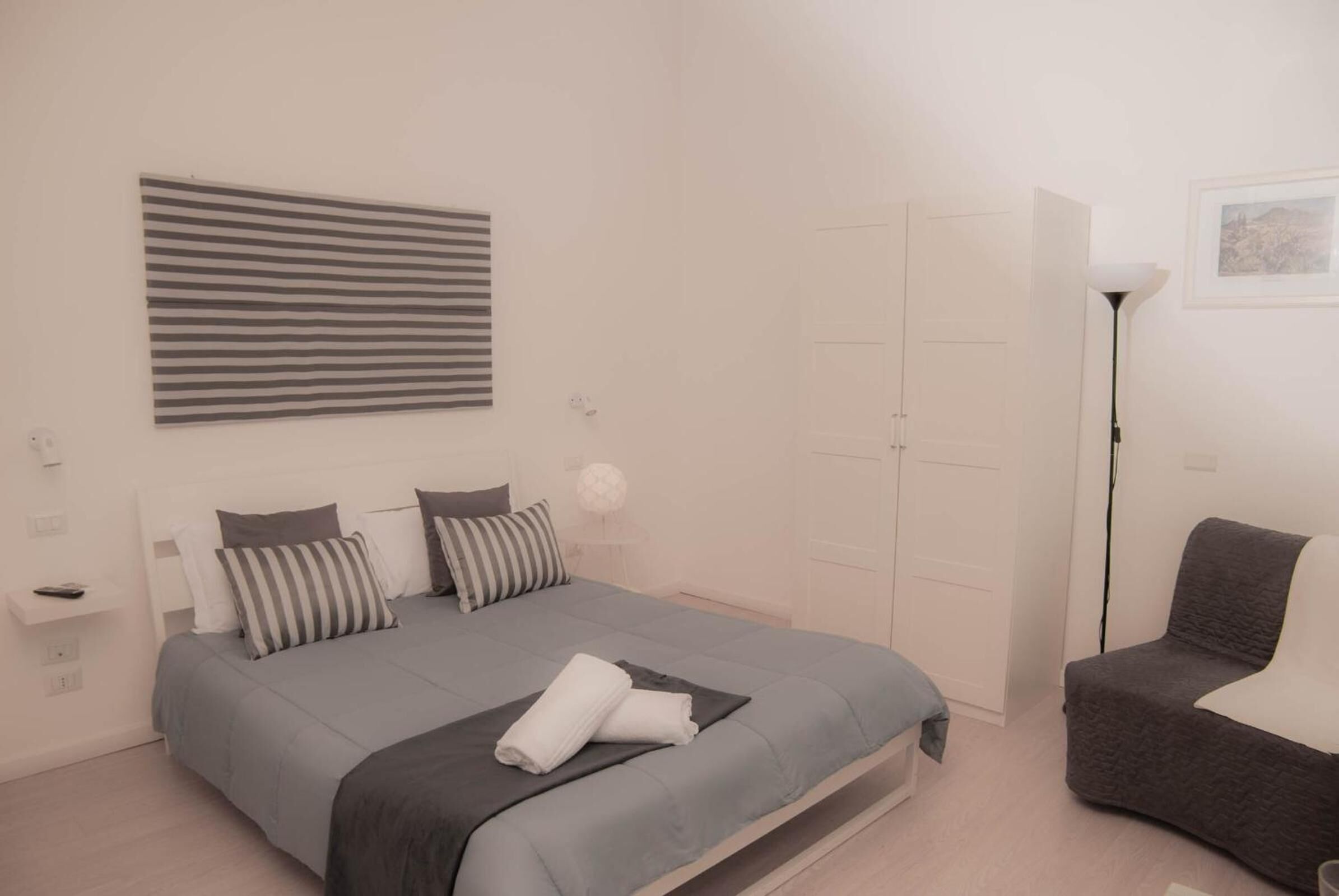 A bedroom with a bed, a bedside table, a lamp, a chair, and a wardrobe.