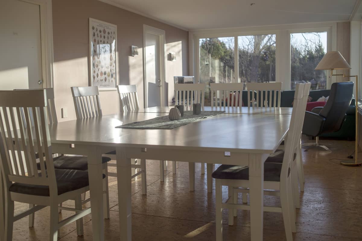 A dining area with a long table and white chairs, a lamp, and a view through large windows.