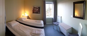 A hotel room with two beds, a desk, a chair, a radiator, a window with curtains, and a wall-mounted mirror. - Klostergården Tautra (Frosta)