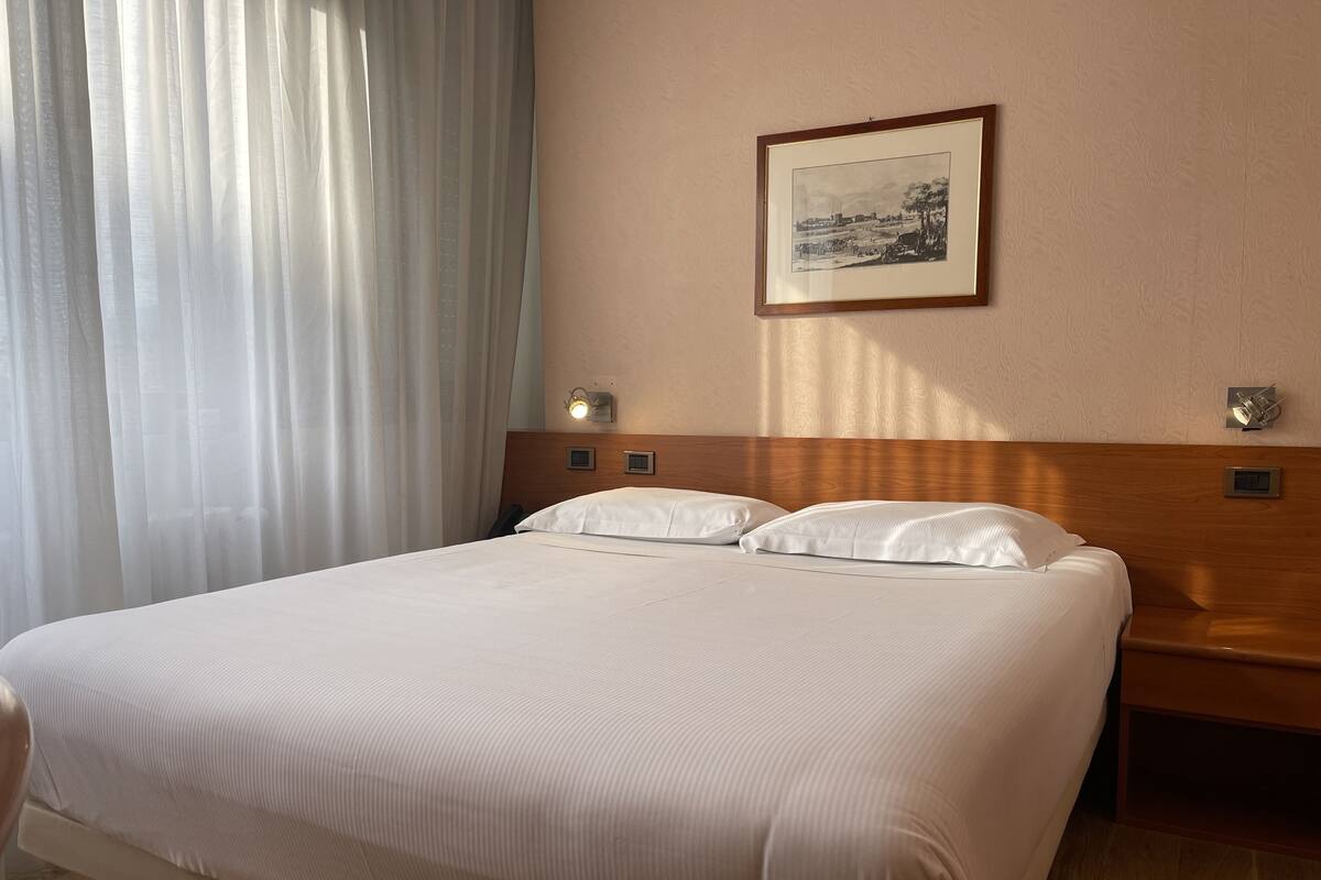 A hotel room with a large bed, a headboard, a nightstand, a framed picture on the wall, and a window with curtains.