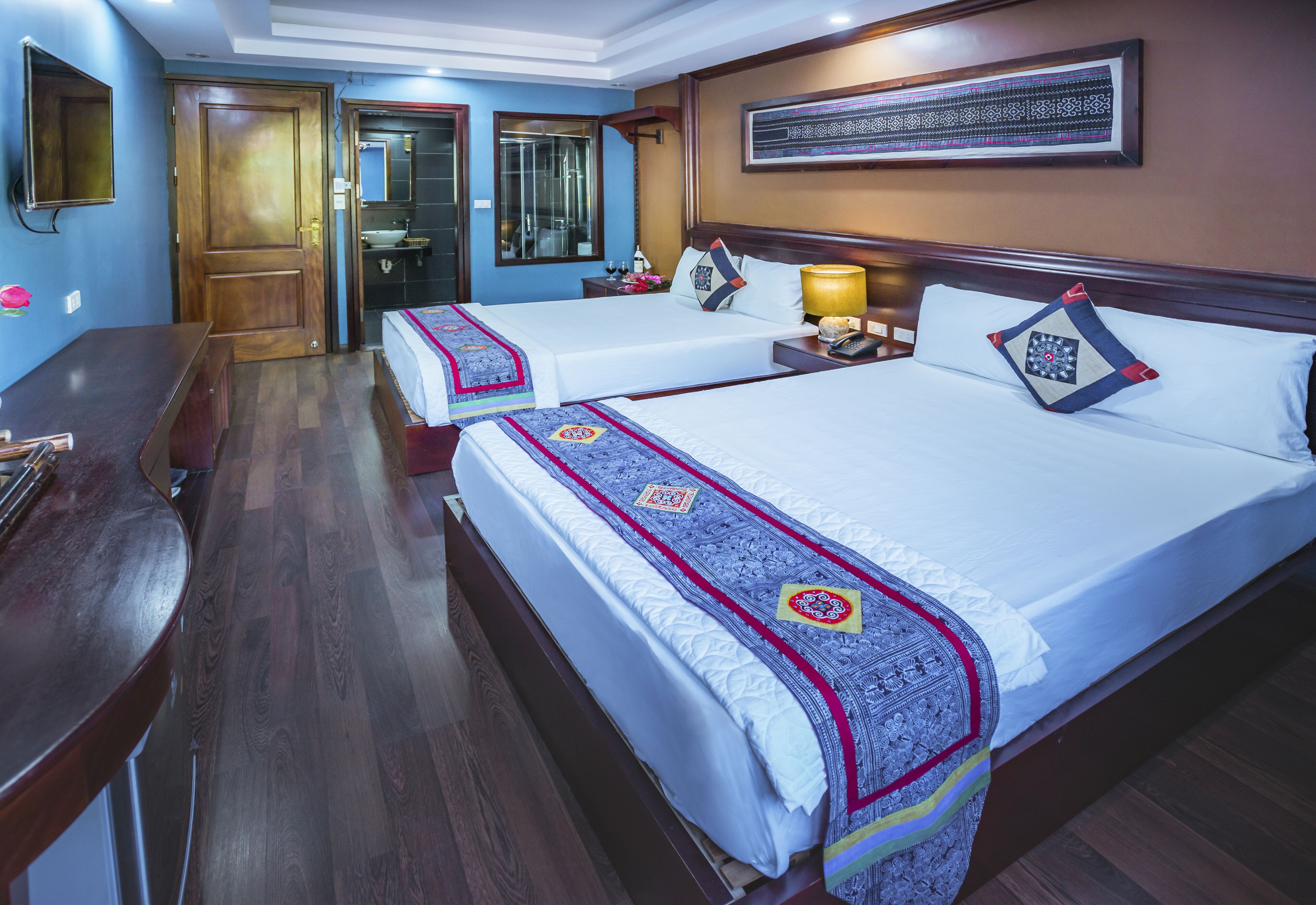 A hotel room with two beds, wooden flooring, a desk, a mirror, a sink, and a framed artwork on the wall.