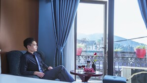 A man sitting on a bed in a hotel room with a balcony view, a table with wine glasses and a bottle, and a potted plant on the balcony. - Sapa Full House Hotel (Sa Pa)
