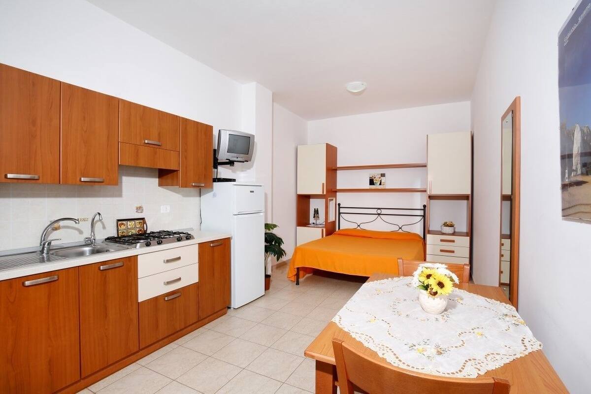 Une chambre d’hôtel compacte, dotée d’une kitchenette, d’un lit recouvert d’une couverture orange, d’une petite table ornée d’un vase de fleurs et d’un miroir au mur.
