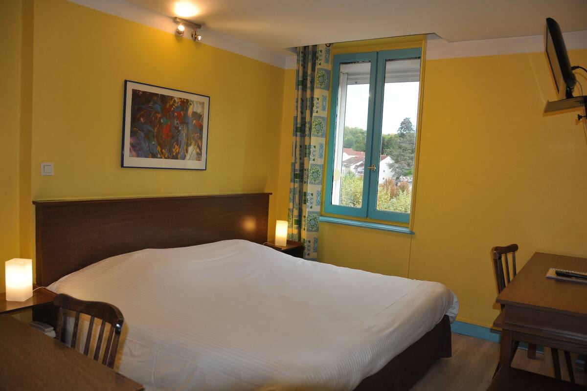 Een hotelkamer met een groot bed, een bureau met stoel, een raam met gordijnen en een schilderij aan de muur.