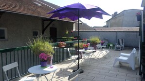 Un área de patio al aire libre con sillas plegables blancas, un paraguas morado y plantas en macetas.