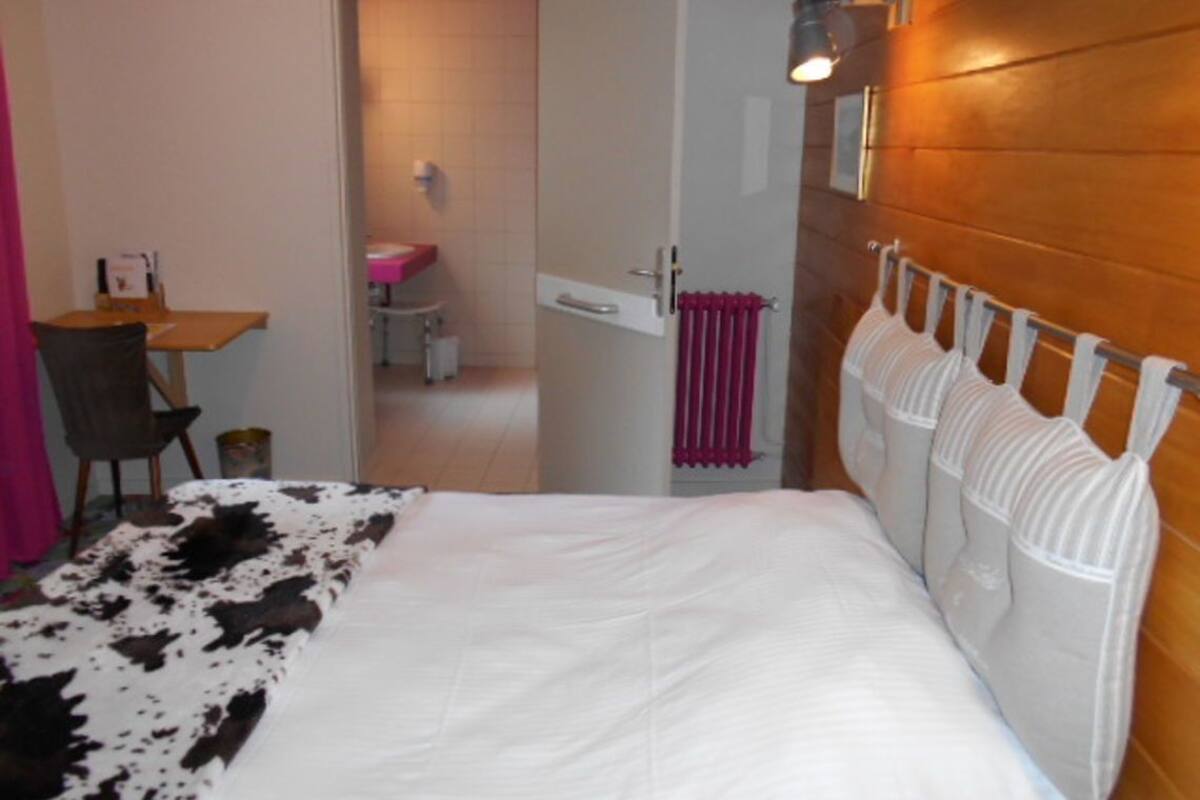 Een slaapkamer met een bed, een houten hoofdeinde, een kleine tafel met stoel, en een zichtbare badkamer door een open deur, plus een roze handdoekhouder.