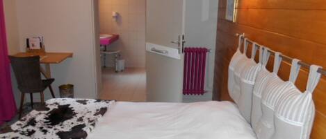 A bedroom with a bed, a wooden headboard, a small table with a chair, and a bathroom visible through an open door.
