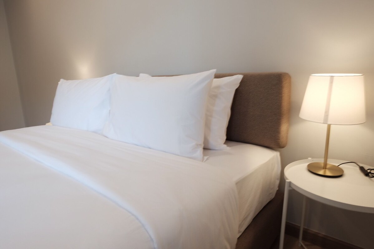 A neatly made bed with white bedding and a single brown pillow. A bedside table with a lamp is visible.