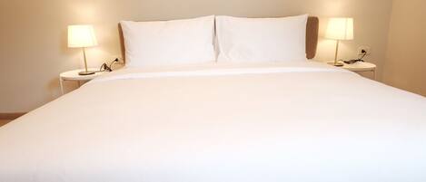 A neatly made bed with white bedding, two bedside lamps, and a plain wall in the background.
