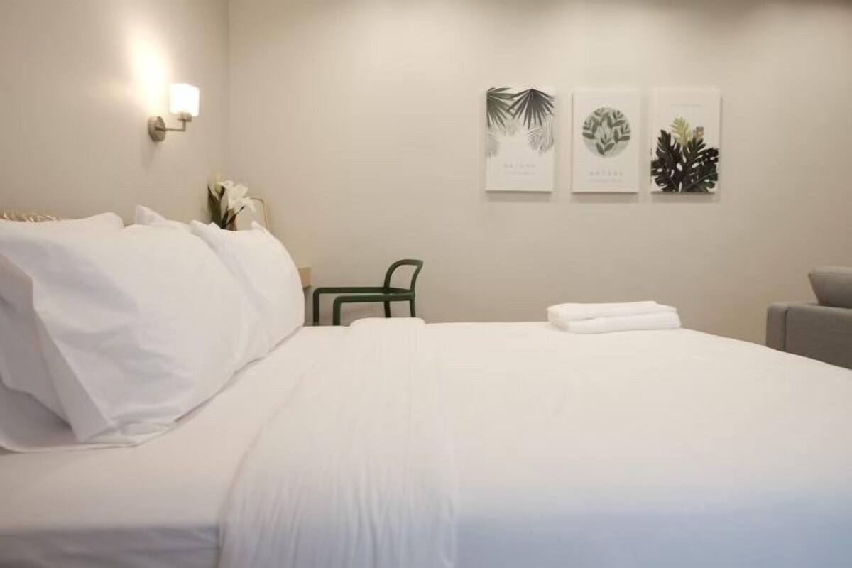 Un lit bien fait, avec du linge de lit blanc, une chaise verte et une petite table sur laquelle est posé un vase de fleurs. Trois reproductions botaniques encadrées ornent le mur.