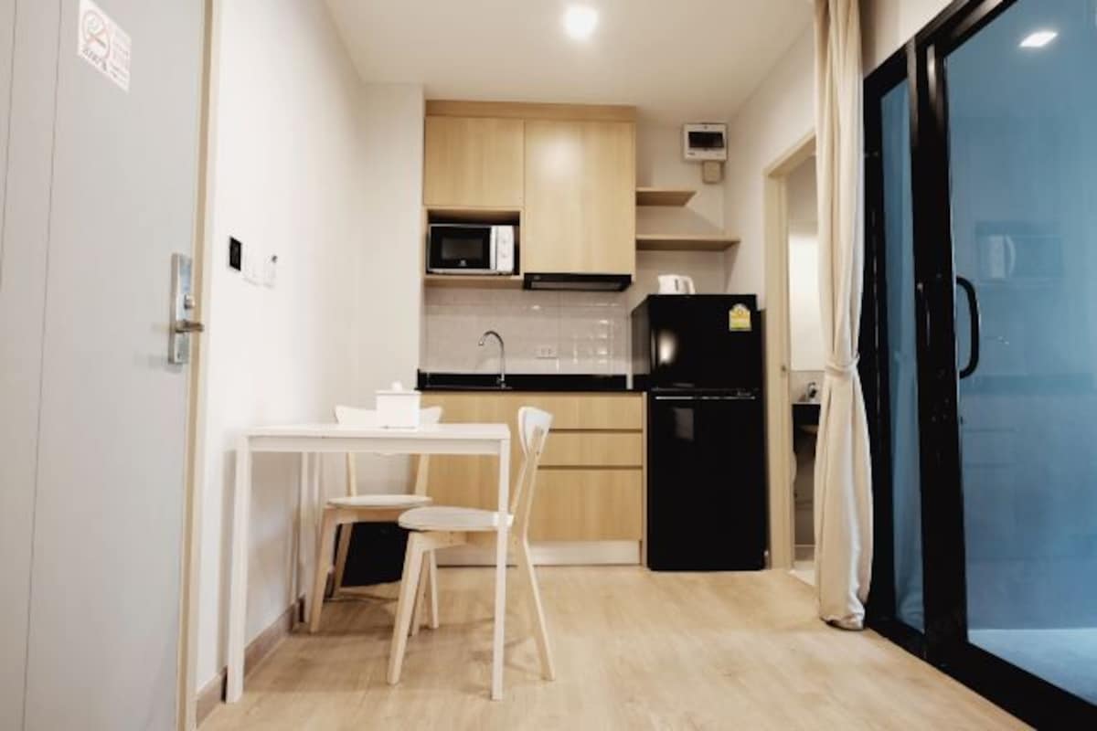 Cocina compacta con armarios de madera, un microondas, un refrigerador negro y una mesa blanca con dos sillas.