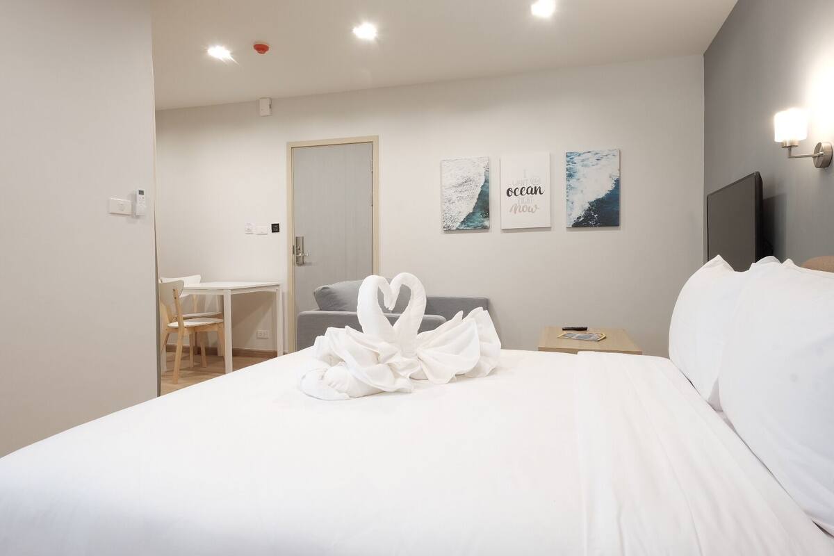 Ein Hotelzimmer mit Bett, Schreibtisch, Stuhl und Fernseher. An den Wänden sind Kunstwerke angebracht und auf dem Bett liegt ein herzförmiges Handtuch.