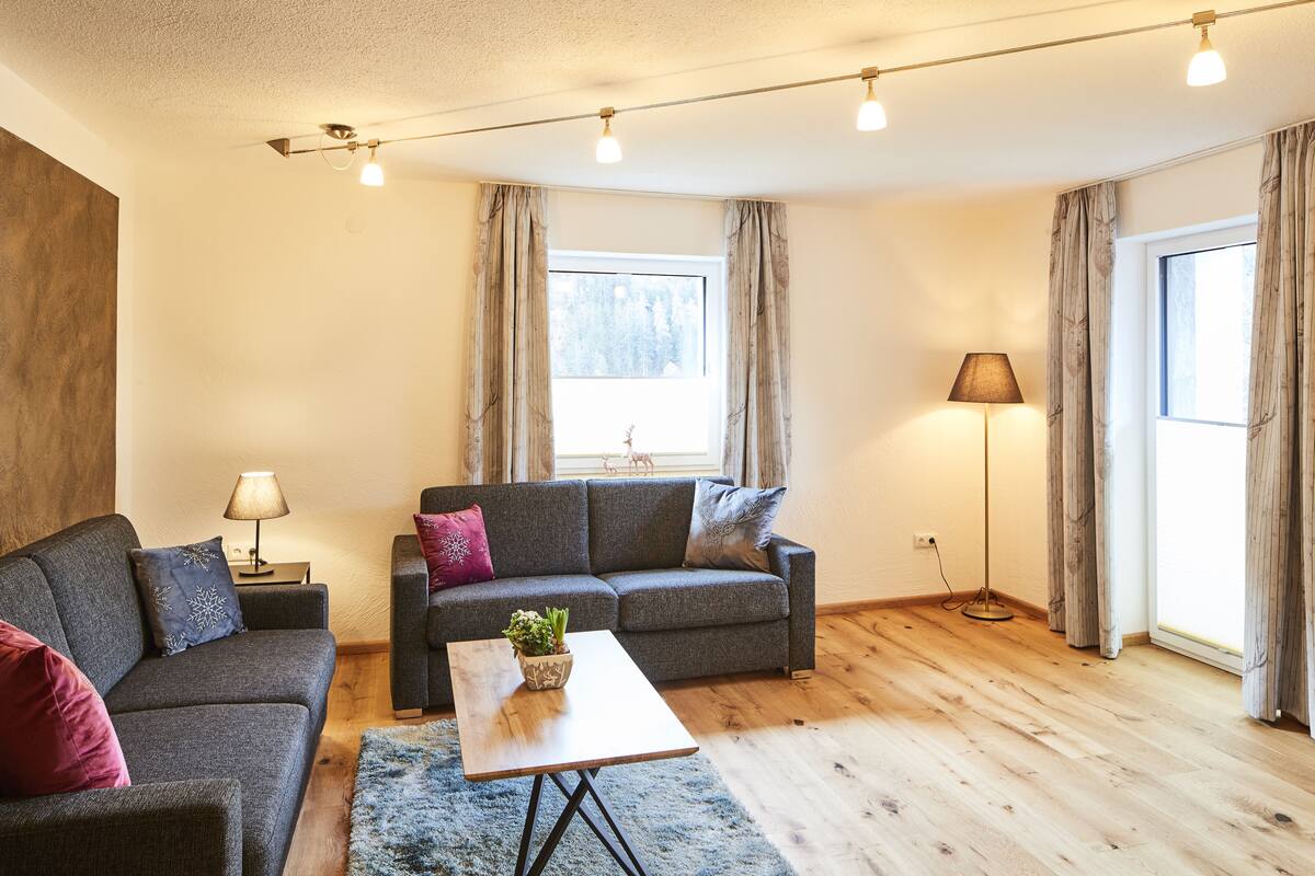A modern living room with a grey sofa, a wooden coffee table, and a blue rug. There are two lamps, a window with curtains, and a view of trees outside.