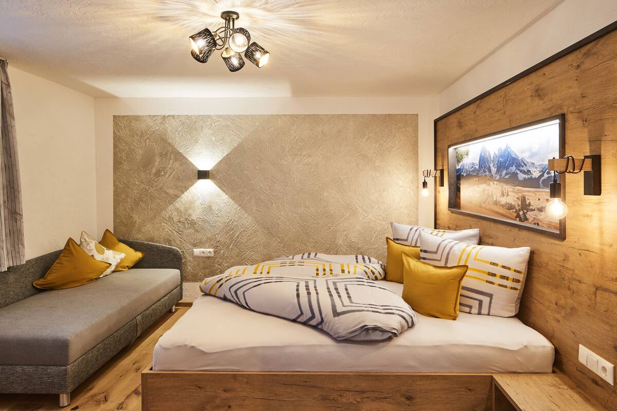 A modern bedroom with a sofa, a bed with patterned bedding, a wall-mounted light, and a framed picture.