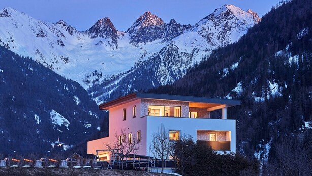 A modern two-story mountain cabin with large windows and a balcony, surrounded by snow-covered peaks and dense forest. - Apartment Naturnah (Kauns)