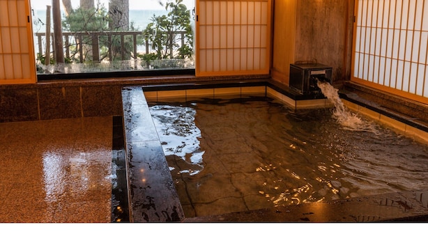 Una tradizionale onsen giapponese con interni in legno, una vasca in pietra e vista su alberi e un pontile in legno all'esterno.